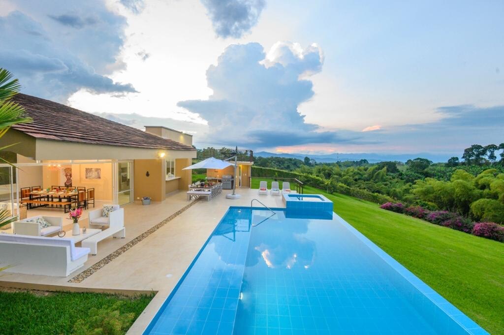 A stunning view of the property captures a large pool reflecting the sky, with an outdoor seating area nearby. The spacious lawn is bordered by lush greenery and colorful flowers, providing a serene backdrop to the elegant house under a softly lit sky.
