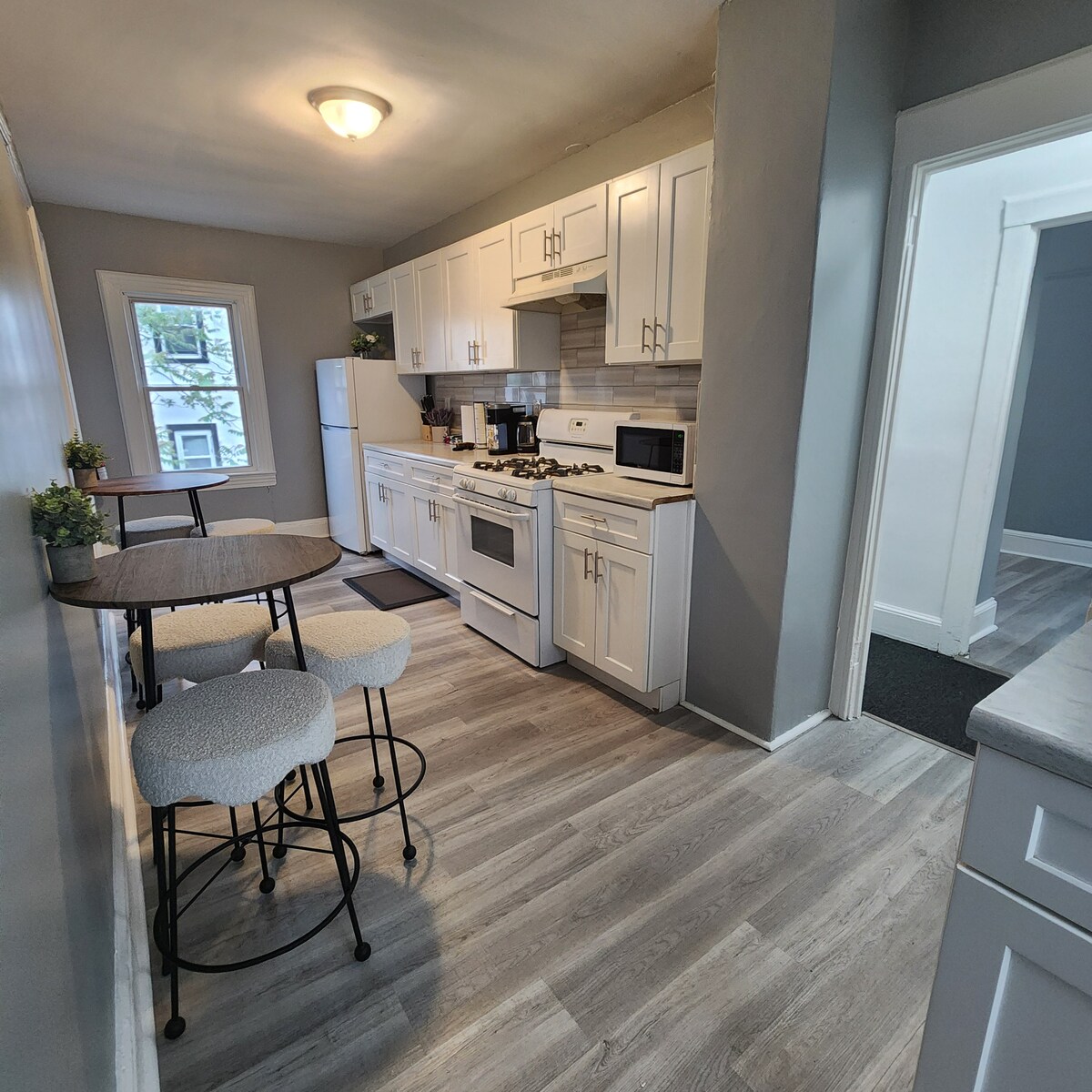 A compact kitchenette is showcased, featuring white cabinetry and modern appliances. A gas stove, microwave, and refrigerator are available, with a small dining table and four upholstered stools. Natural light enters through a window, enhancing the space's brightness and functionality.