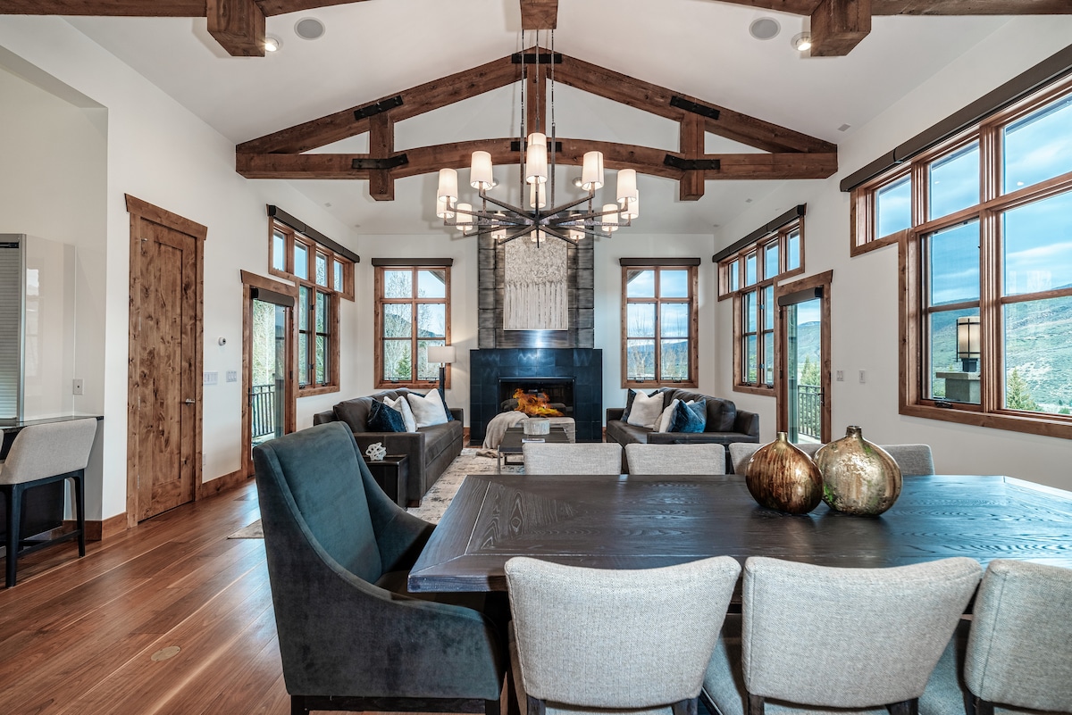 The living area features towering windows that allow natural light to fill the space, showcasing mountain views. A plush sectional sofa is positioned near a stone fireplace, while a dining table sits nearby, accompanied by cozy seating options. Exposed wooden beams highlight the room's design.