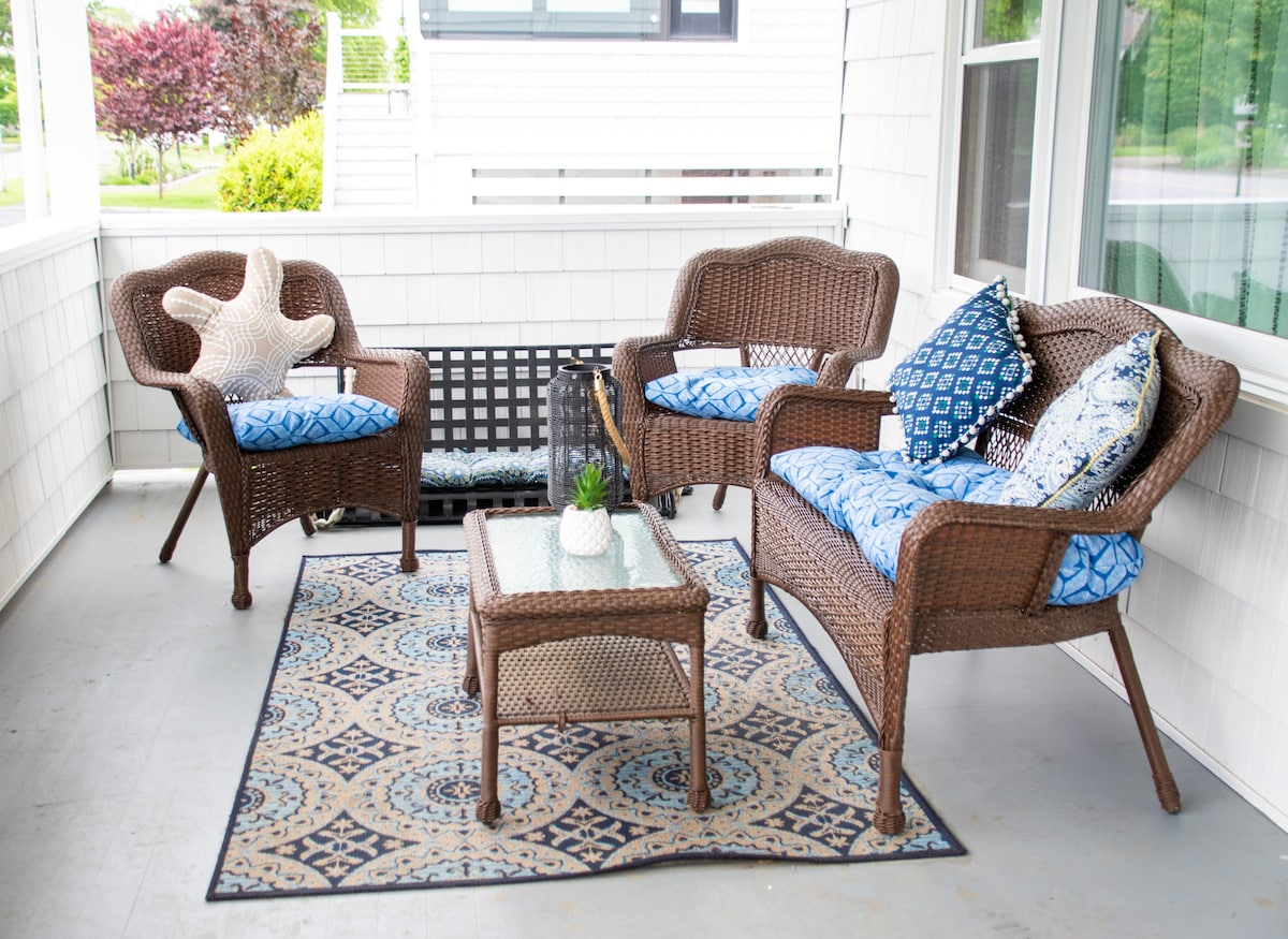 An inviting front porch area features a set of comfortable wicker chairs with decorative blue cushions, accompanied by a matching coffee table. The space is anchored by an intricately patterned rug, while a small plant adds a touch of greenery.