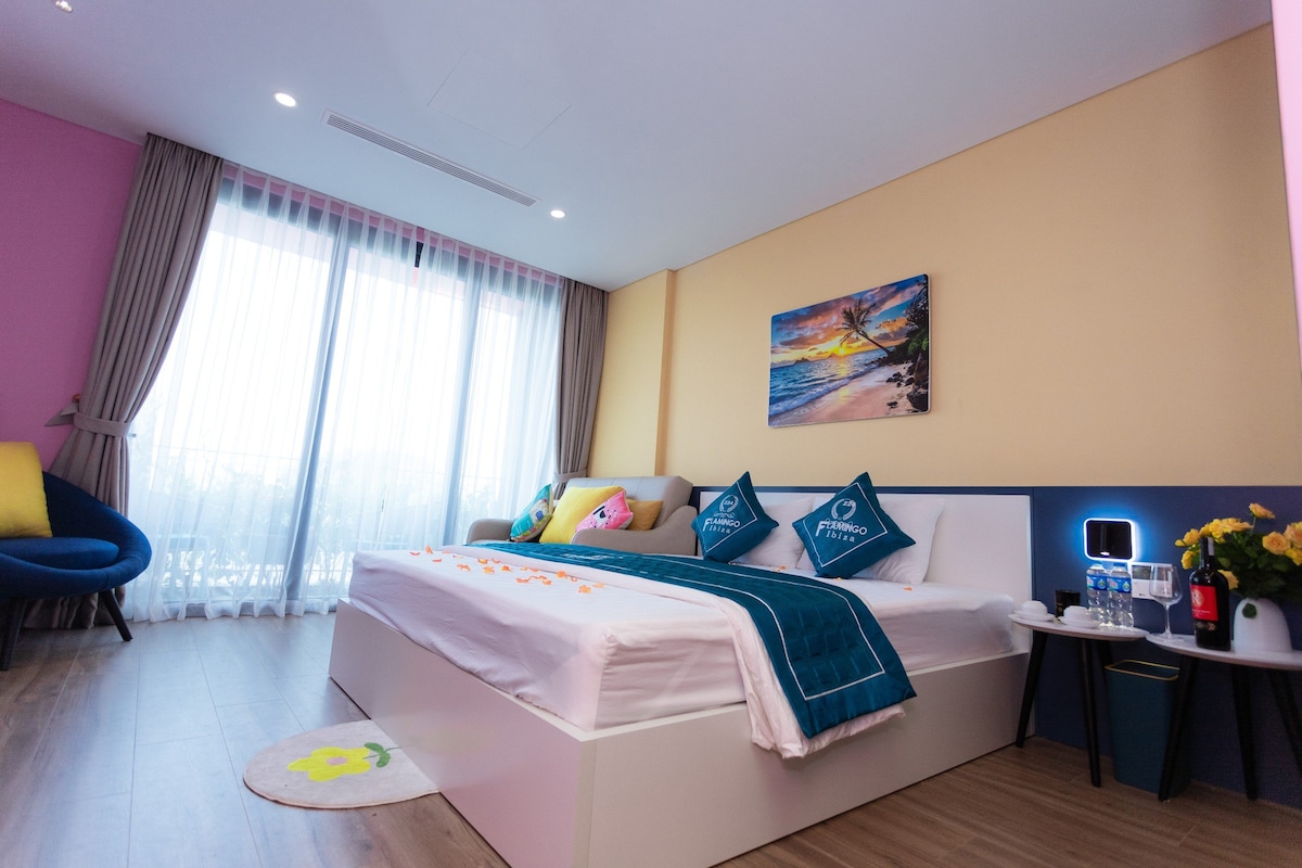 A bright sleeping area features a spacious bed with decorative pillows on a light-colored platform. A cozy sofa sits beside the bed, complemented by a small table and a vibrant wall art piece. Large windows provide natural light, enhanced by soft curtains.