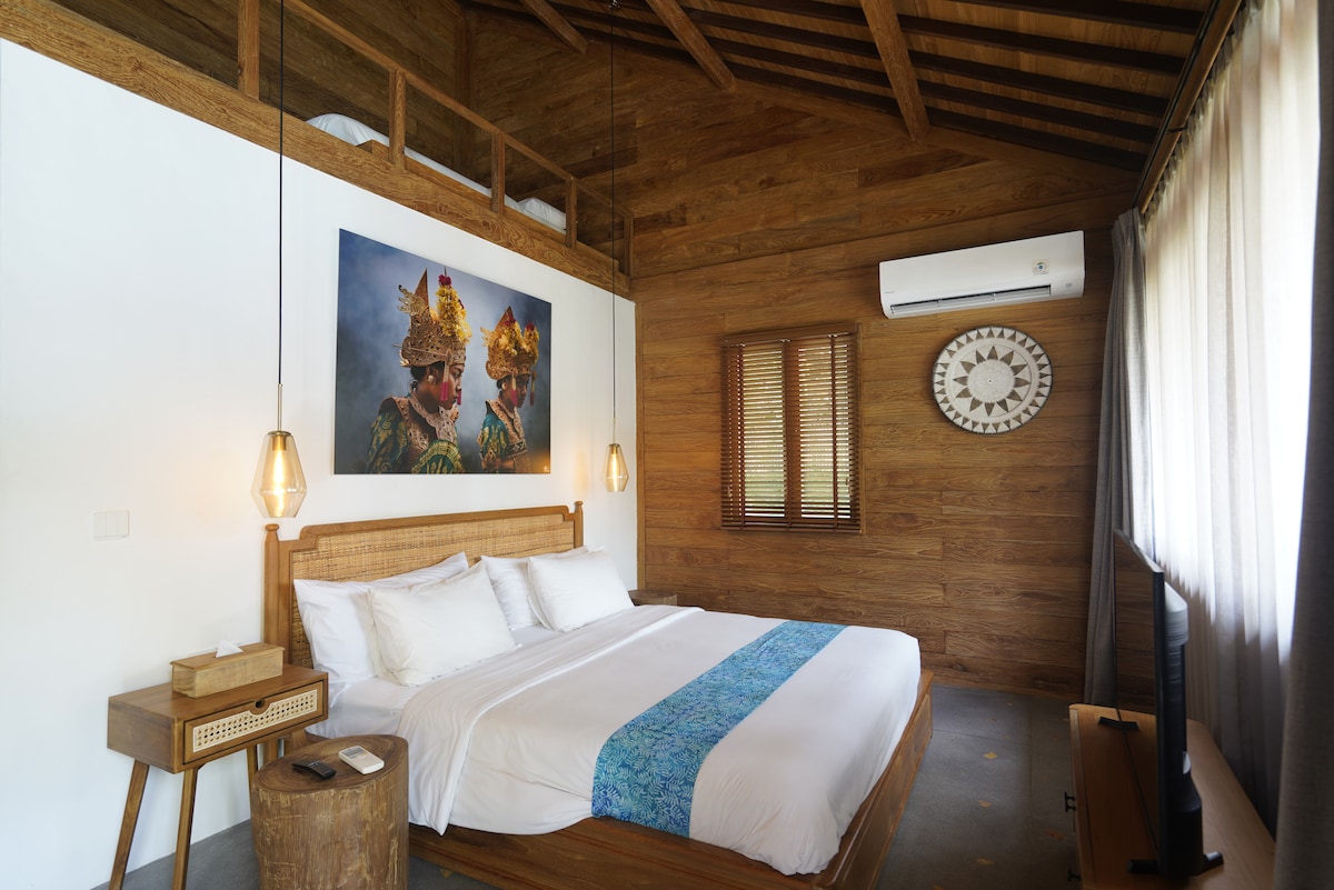 The cozy bedroom features a spacious bed adorned with a white comforter and a decorative blue accent. Natural wood elements and warm lighting create an inviting ambiance. A large artwork depicting traditional figures is displayed above the bed, complemented by a wall clock and a smart TV.