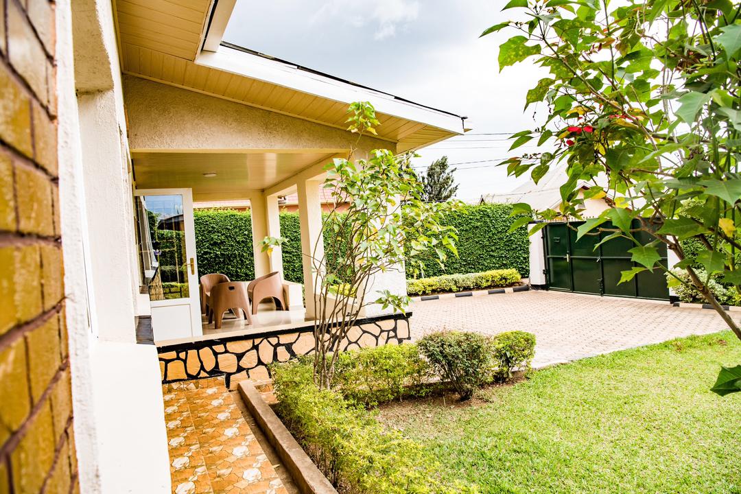 A spacious veranda is visible, supported by tall columns. Comfortable outdoor seating is arranged, surrounded by lush green foliage. Neatly manicured grass leads to a driveway, while tall hedges create a sense of privacy. A well-kept garden enhances the inviting outdoor space.