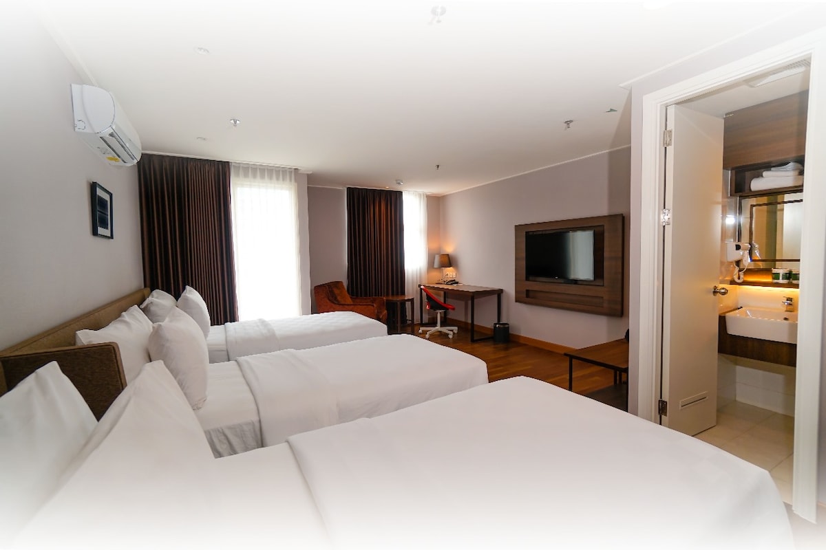 A well-appointed room features three spacious beds arranged for comfort. A working desk and cozy armchair are positioned near the window, allowing plenty of natural light. An LED TV is mounted on the wall, with an adjoining bathroom visible in the background.