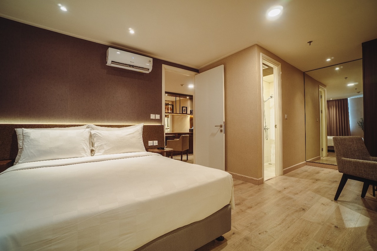 A spacious room is featured, showcasing a king-sized bed with a simple white headboard. A modern air conditioning unit is mounted above the bed. The room is illuminated with soft lighting, and a doorway leads to a well-lit bathroom. A comfortable chair is positioned nearby.