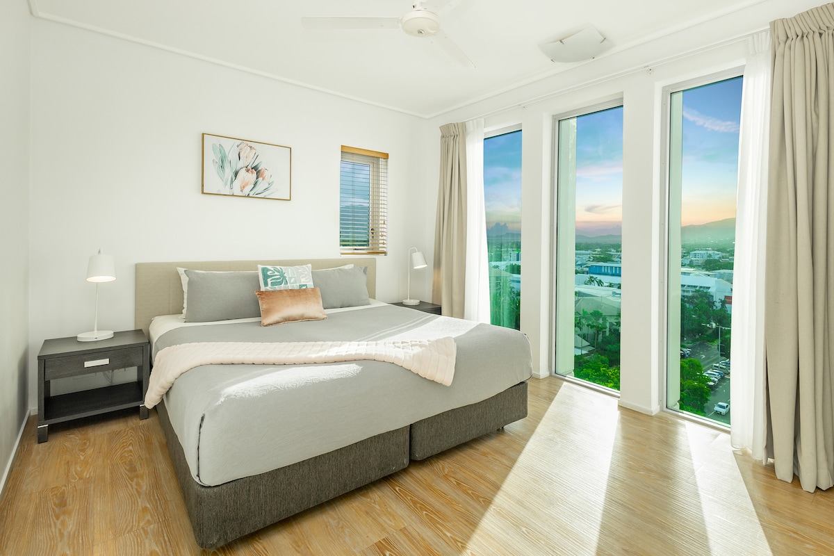A spacious bedroom features a king-sized bed dressed in light gray bedding, positioned against a light wall. Floor-to-ceiling windows offer views of Cairns city, with a sunset glow reflected in the glass. Nightstands with lamps are placed on either side of the bed.