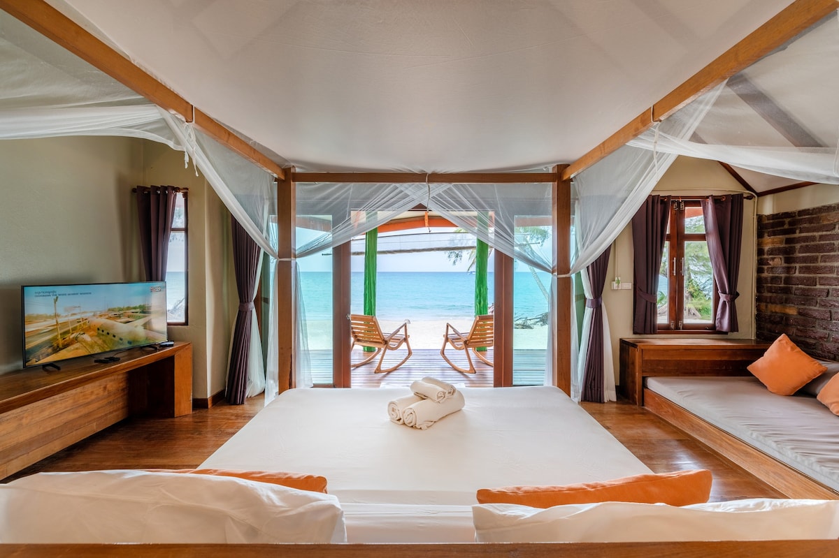 A spacious sleeping area features a large bed with rolled towels positioned at the center. Two wooden chairs are visible through the open doors, overlooking the sea, while a television is placed on a wooden stand and natural light filters in through nearby windows.