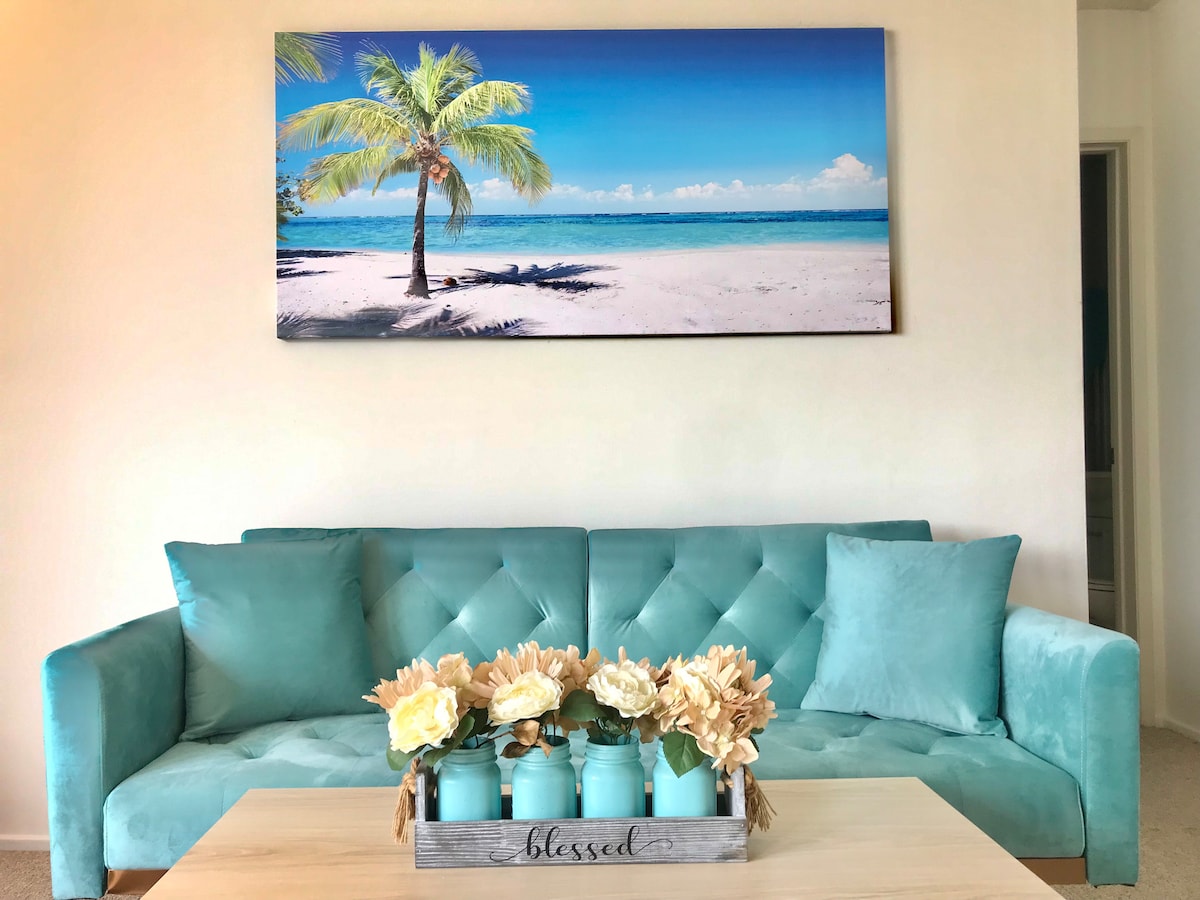 A comfortable aqua sofa is presented in a inviting living room, accompanied by a light wooden coffee table. Above the sofa, a landscape photograph of a serene beach with a palm tree overlooks the space. A decorative tray holds flowers, enhancing the relaxed ambiance.