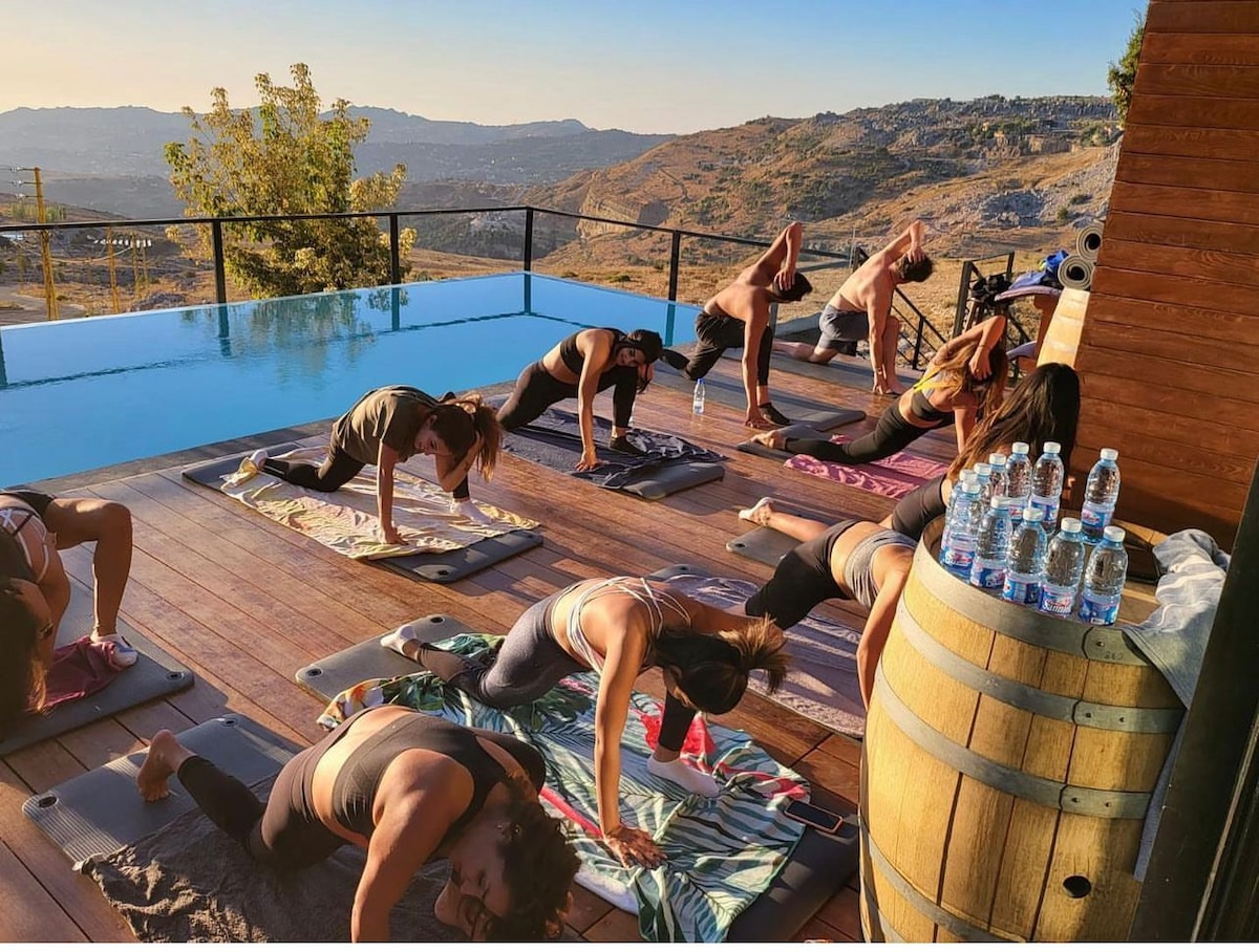 A spacious outdoor deck overlooks rolling hills, featuring a group engaged in a yoga class on mats. A tranquil infinity pool reflects the sky, and water bottles are neatly arranged on a nearby barrel, providing hydration for participants.