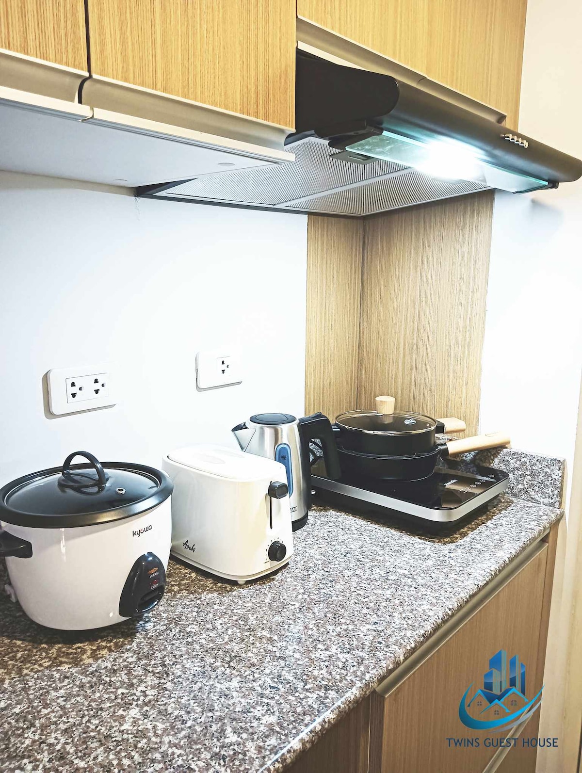 A well-equipped kitchen features modern appliances, including a rice cooker, kettle, and a frying pan on a cooktop. A granite countertop provides ample workspace, with a light wood cabinet above for storage.
