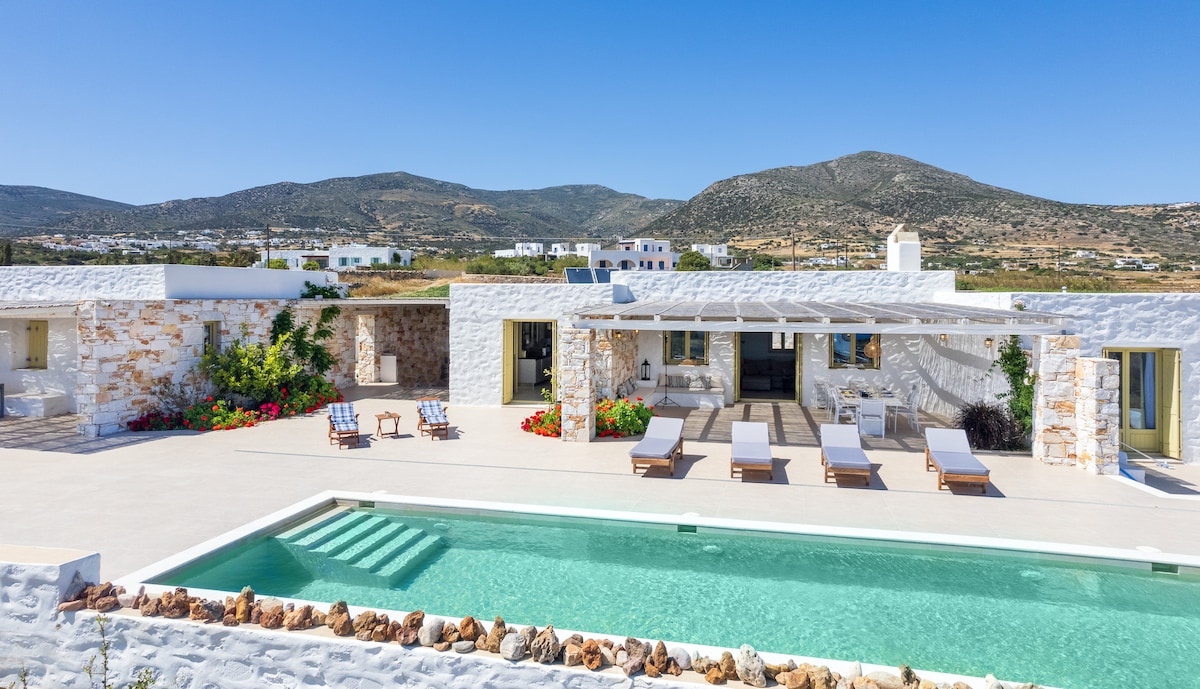 A newly built beach villa features a private swimming pool with steps leading into the water. Lounge chairs are arranged around the pool area, accompanied by landscaped gardens. The background displays rolling hills under a clear blue sky, creating a serene and spacious atmosphere.