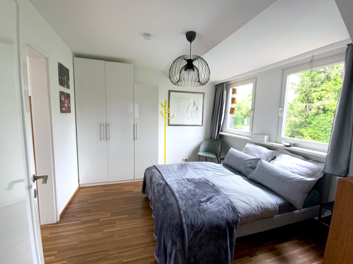The bedroom features a spacious bed dressed in soft linens. A large wardrobe occupies one wall, while a bright yellow coat rack adds color. Natural light flows through the windows, offering views of the surrounding gardens. The light wood flooring contributes to the room's overall brightness.