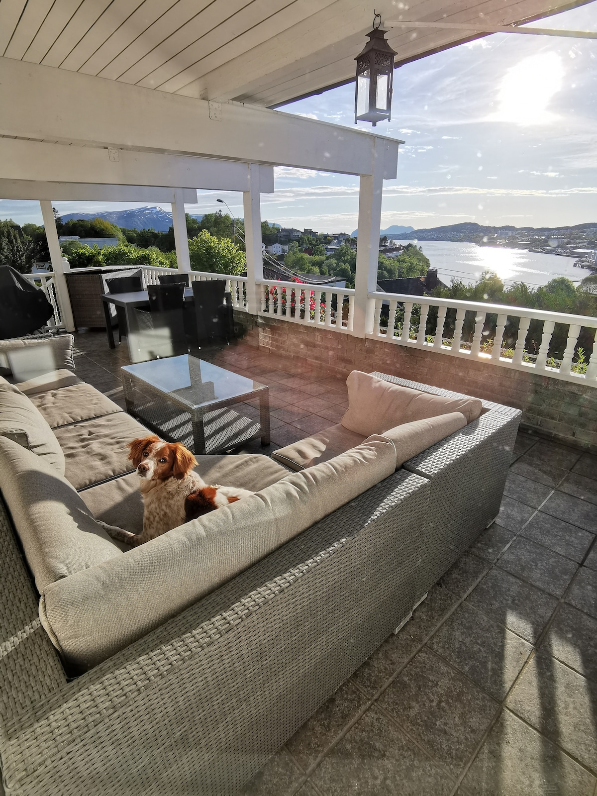 A spacious outdoor seating area features a comfortable sectional sofa in neutral tones, complemented by a glass coffee table. Scenic views of the water and surrounding landscape are visible in the background, enhanced by sunlight reflecting off the surface.