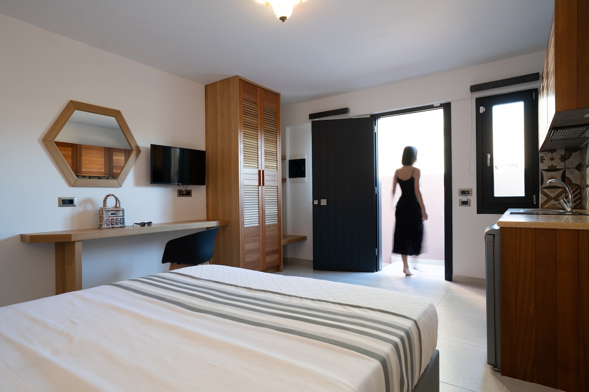 A bright studio space features a spacious bed with a grey-striped blanket. A handmade wooden mirror and closet are present. A door leads to an outdoor area, with natural light filling the room. A small kitchenette is visible, accompanied by modern appliances.