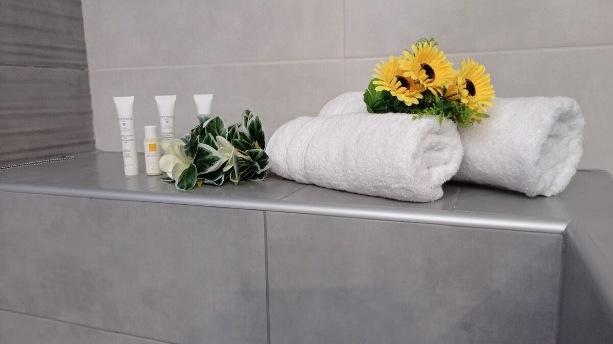 A clean and inviting bathroom counter displays neatly rolled white towels alongside small tubes of toiletries. A delicate arrangement of sunflowers and greenery adds a touch of decor to the space, creating a fresh and welcoming ambiance.