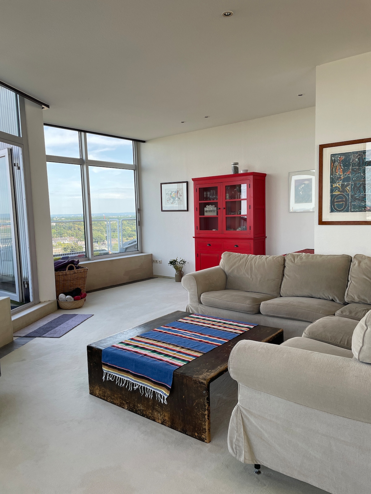 A bright living area features large windows offering sweeping views of the surroundings. A comfortable light-colored sofa is arranged with a striped throw. A rustic wooden coffee table is positioned in front, complemented by an eye-catching red cabinet against the wall.