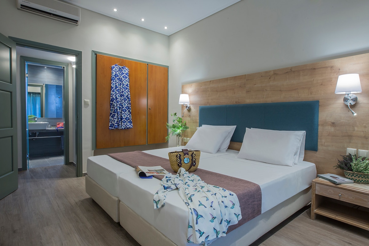 The suite features two spacious beds with crisp white linens and a muted color palette. A decorative throw and items like books are placed on the bed. Soft lighting from wall sconces illuminates the room, while a doorway leads to a bathroom, enhancing accessibility.