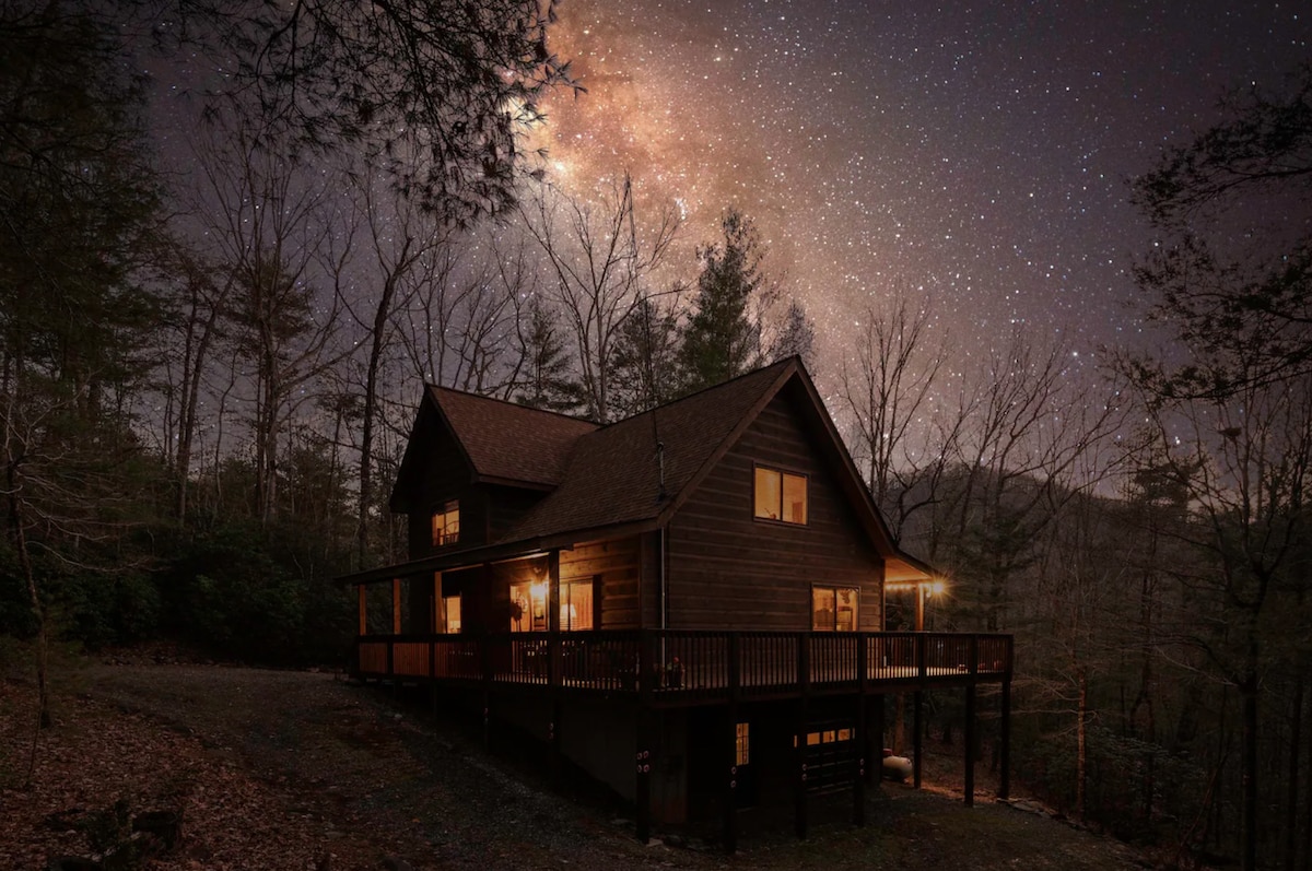 A cabin is nestled among tall trees, illuminated warmly by outdoor lighting, showcasing a wraparound deck. Above, a clear night sky filled with stars and the Milky Way adds to the serene atmosphere, highlighting the peacefulness of the surrounding nature.