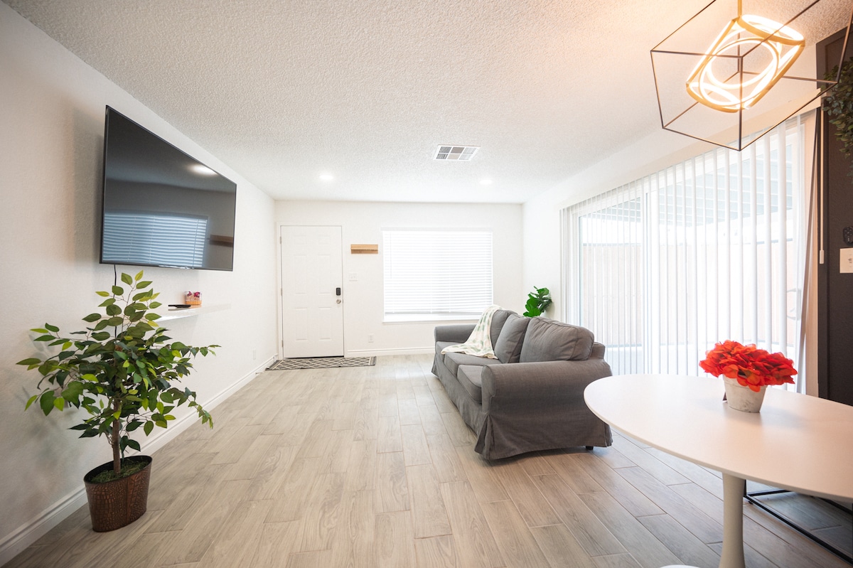 A living area offers a spacious couch facing a wall-mounted TV. A round table with chairs is present near a sliding glass door that opens to a patio. Natural light enters through the window, complemented by a small plant in a pot.