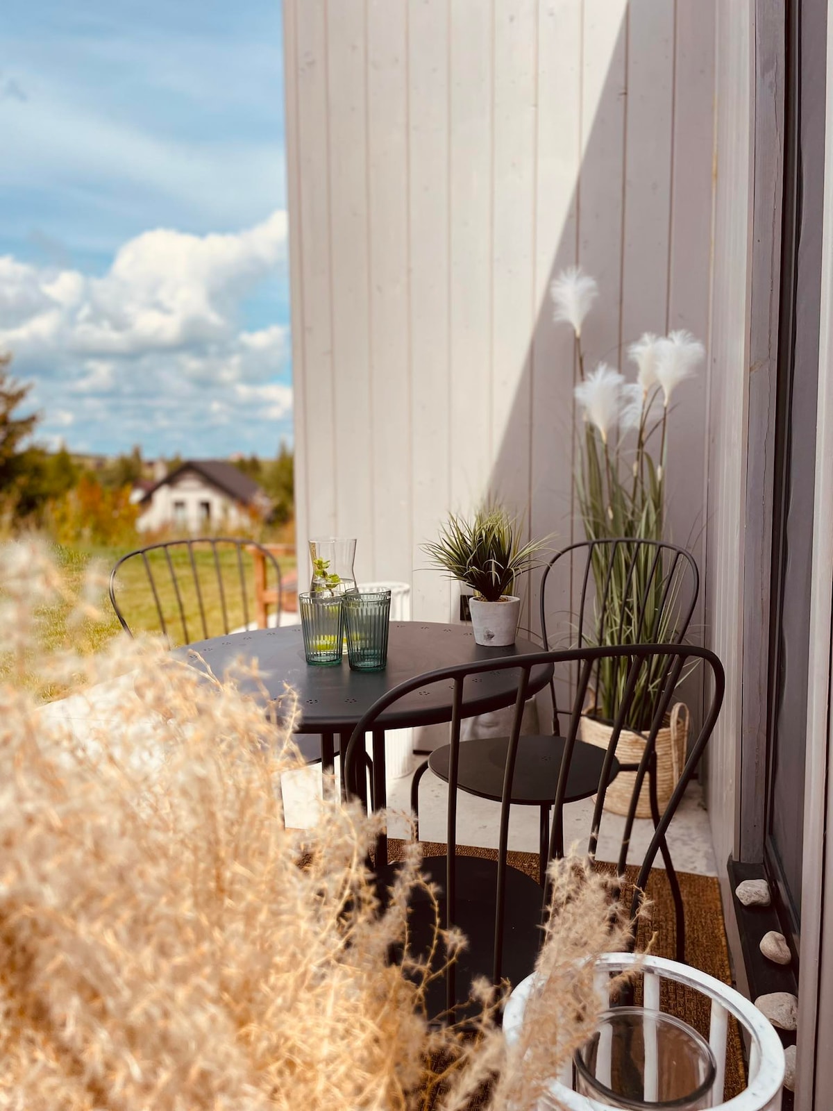 A small outdoor seating area is showcased, featuring a round table accompanied by two black chairs. Decorative elements include a green plant in a pot and clear glasses on the table. Tall grasses frame the scene, enhancing the natural setting with a backdrop of blue skies and distant greenery.