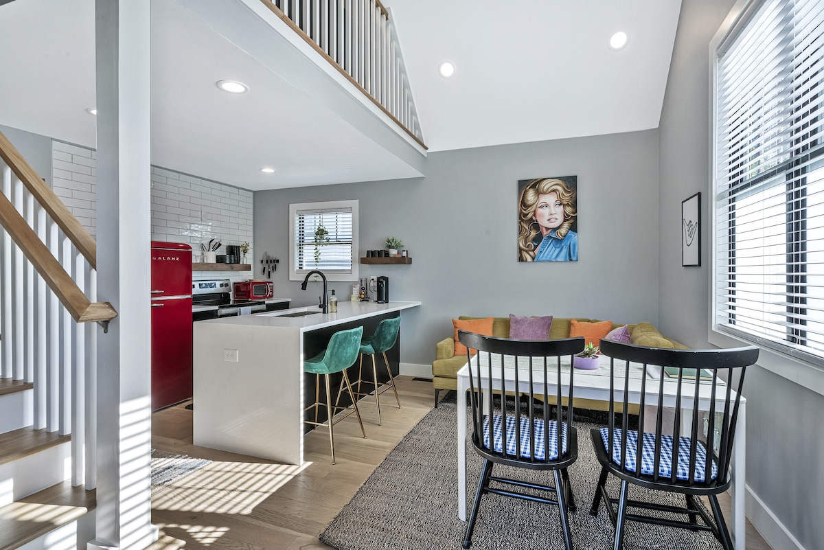 A modern kitchen and living area are visible, showcasing a red retro refrigerator, white countertops, and teal bar stools. A cozy couch with colorful cushions complements a dining table with black chairs. Sunlight enters through large windows, illuminating the space.
