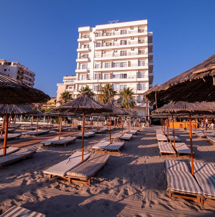 A modern multi-story building is set against a clear blue sky, with several straw umbrellas providing shade over sun loungers arranged neatly on sandy ground. Palm trees add a touch of greenery, complementing the beachside environment.