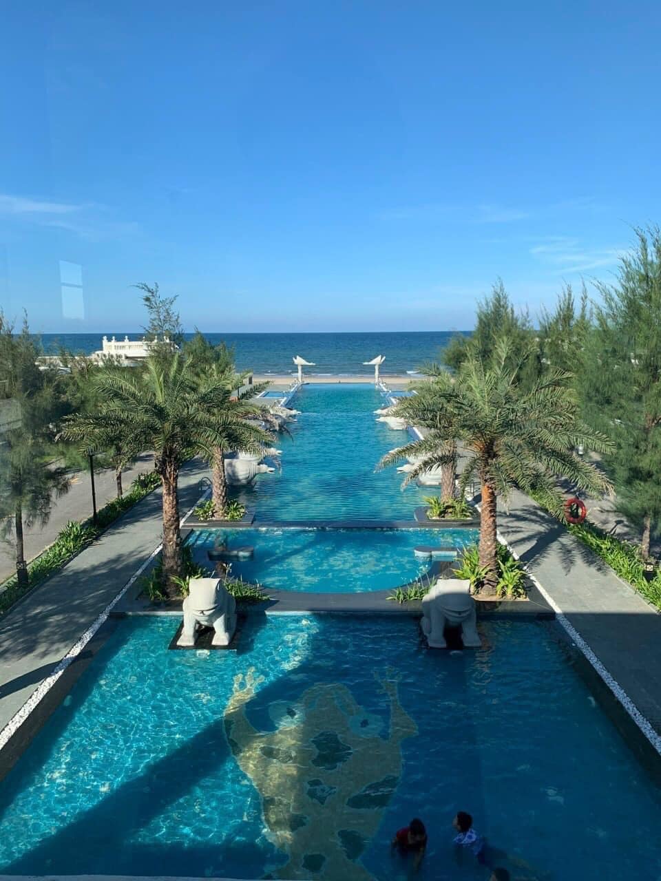 A serene outdoor pool area is flanked by palm trees, leading towards the ocean. The water features a vibrant blue hue, with comfortable loungers positioned along the sides. In the distance, the beach and clear sky create a tranquil backdrop.