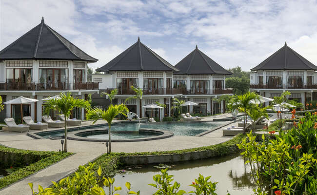 A serene villa complex is showcased, featuring elegant architecture with pointed roofs. Surrounding the outdoor public swimming pool are lounge chairs and tropical plants, creating an inviting atmosphere. The building’s balconies overlook the pool and landscaped gardens, complemented by a tranquil water feature.