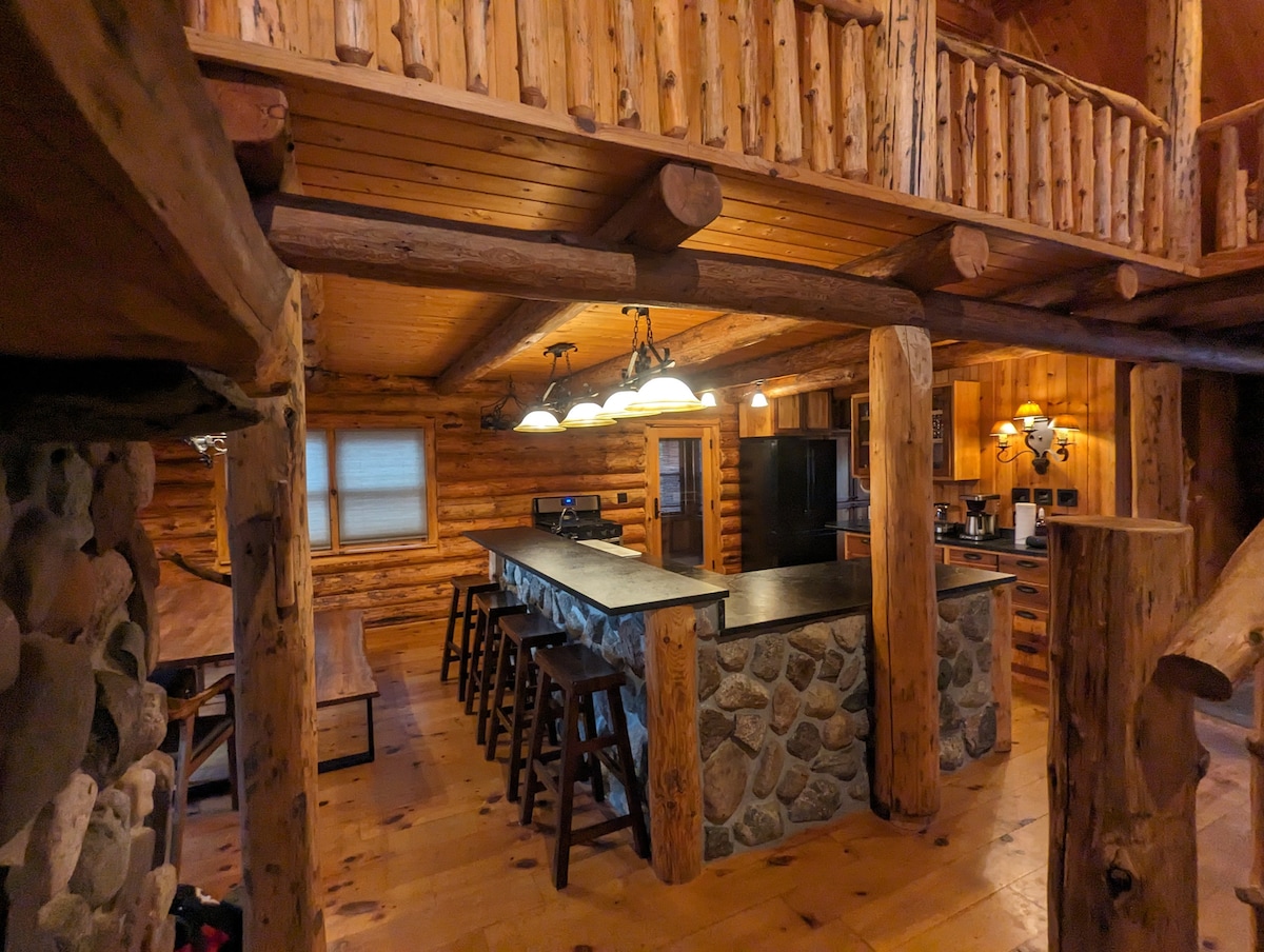 The kitchen area features a large stone island with bar seating, surrounded by wooden beams and rustic decor. Modern appliances are present, and natural light filters through a window, illuminating the inviting and functional space.