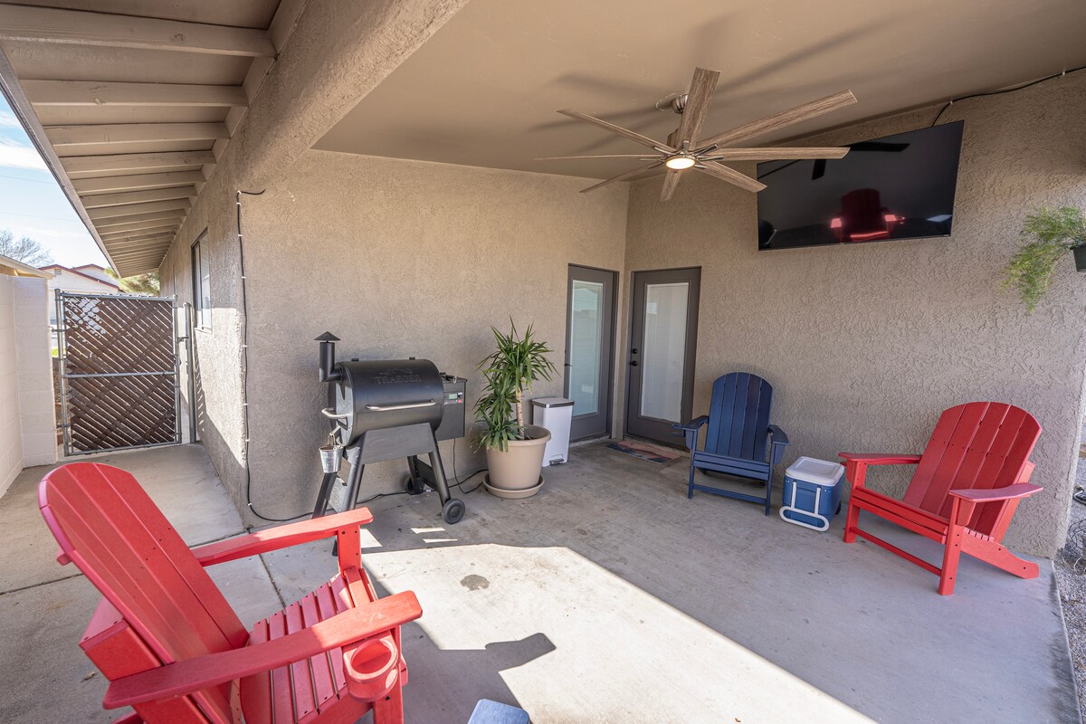 A covered patio area includes two red adirondack chairs and a blue one, inviting relaxation. A ceiling fan provides airflow, while a large television is mounted on the wall. A grill and potted plant add to the outdoor comfort, enhancing the space for gatherings.