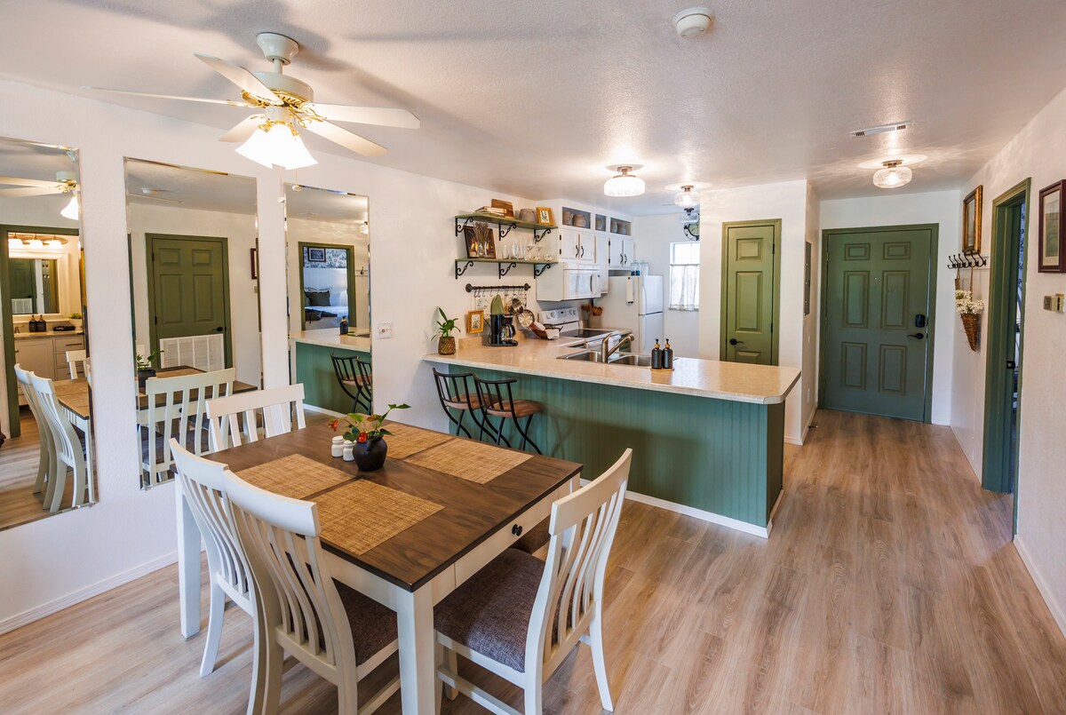 A bright dining area features a wooden table with four chairs, complemented by a small plant centerpiece. Open shelves and decorative items are visible in the background, along with a spacious kitchen area. Light-colored walls and a neutral floor enhance the overall brightness.