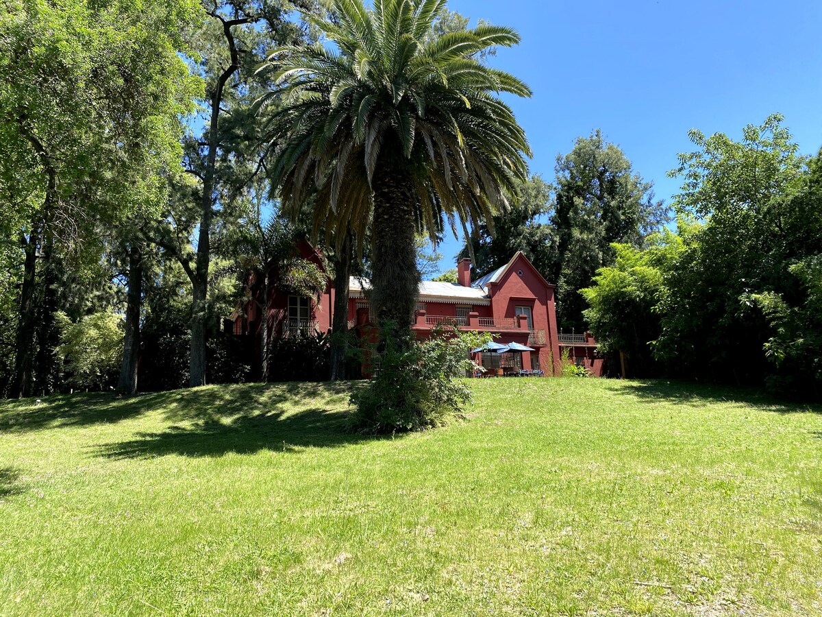 A historic house, characterized by its reddish facade, is set against a backdrop of lush greenery. Towering palm trees and other foliage surround the expansive grassy area in the foreground, providing a serene and natural atmosphere.