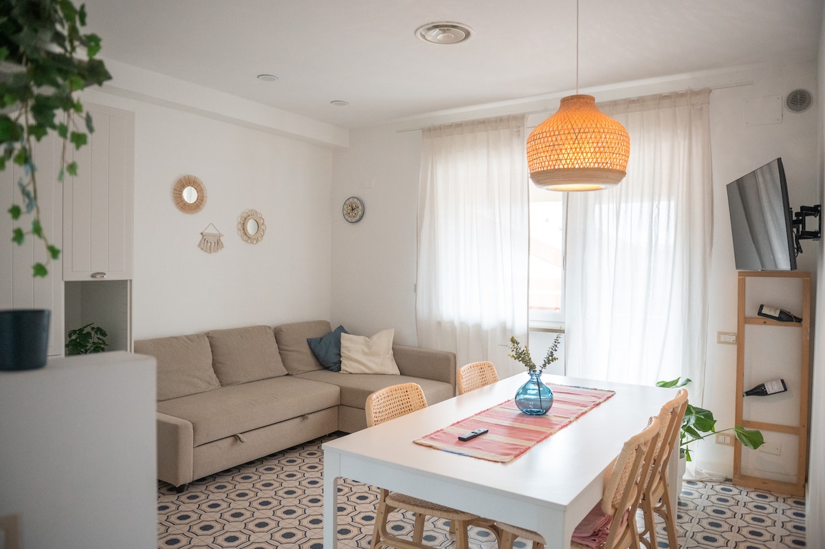 An open-plan living area features a light-colored sectional sofa, a white dining table with wooden chairs, and a large window adorned with sheer curtains. A woven pendant light hangs above the table, while decorative wall pieces add subtle interest to the room.
