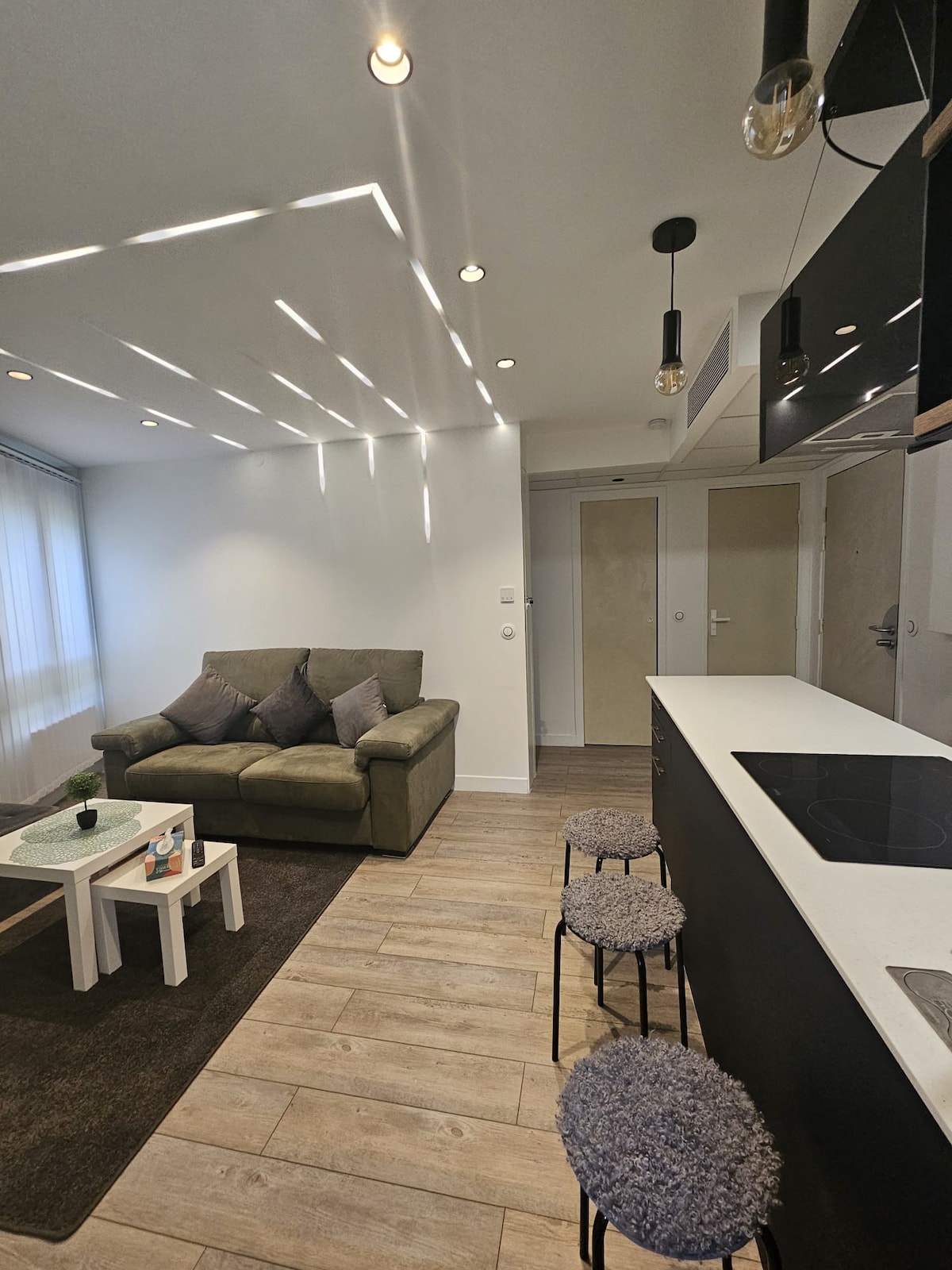 A living space features a cozy green sofa adorned with four decorative pillows. A small coffee table sits in front, and two stylish stools are placed at the kitchen counter. Soft lighting highlights the room's modern design with warm, neutral tones.
