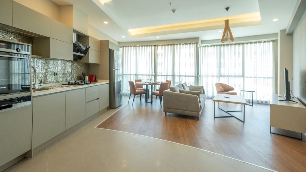 An open-plan living area displays a modern kitchen with sleek cabinetry and stainless steel appliances. Comfortable seating arrangements are provided with a light-colored sofa and two armchairs. Large windows allow ample natural light to fill the space, complemented by minimalist decor.