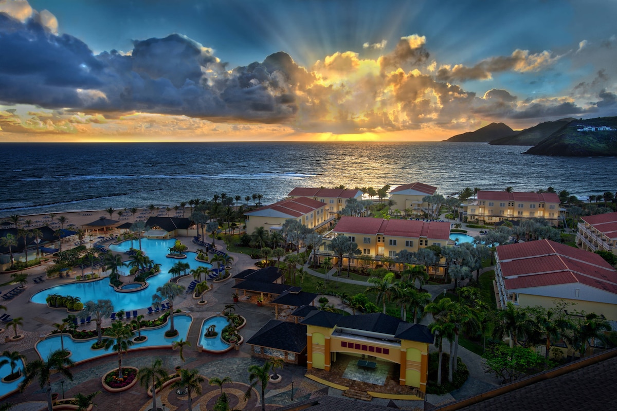 A panoramic view captures the resort's layout alongside the ocean at sunset, with vibrant colors illuminating the sky. The scene showcases multiple pools surrounded by lush greenery and charming building designs that complement the natural surroundings.