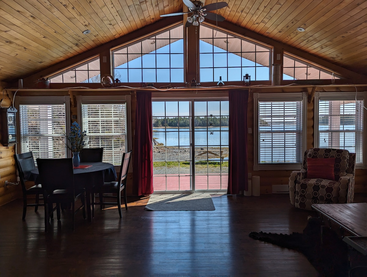 A bright interior showcases a dining area with a table and chairs beside expansive glass doors. Natural light fills the space, highlighting the wooden ceiling and floors. Outside, views of the water and nearby nature can be seen through the large windows.