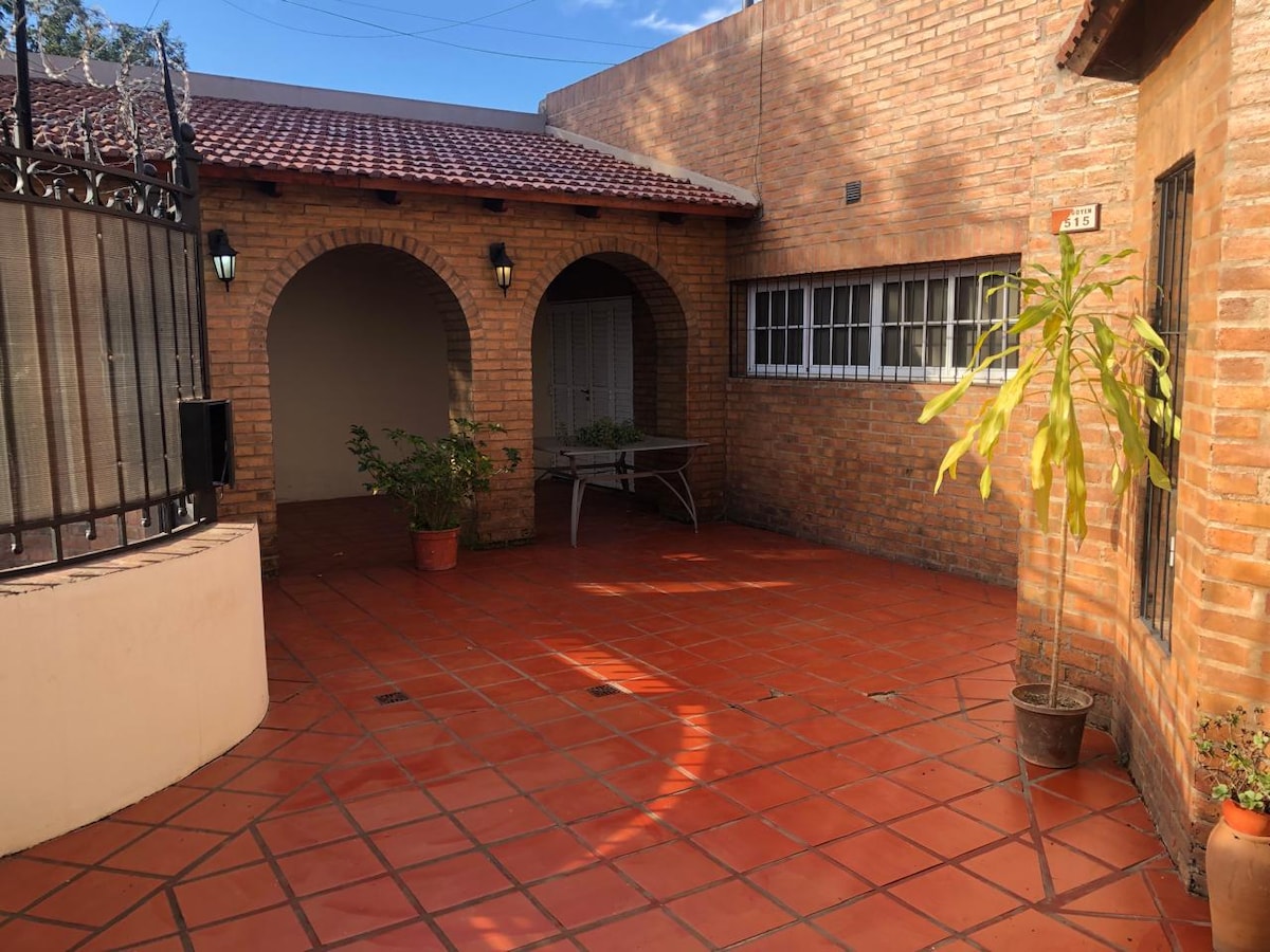 A private outdoor space features terracotta tile flooring with several potted plants placed thoughtfully around. An arched entrance leads to a sheltered area with seating. Natural light illuminates the space, highlighting the warm brick walls and the surrounding greenery.