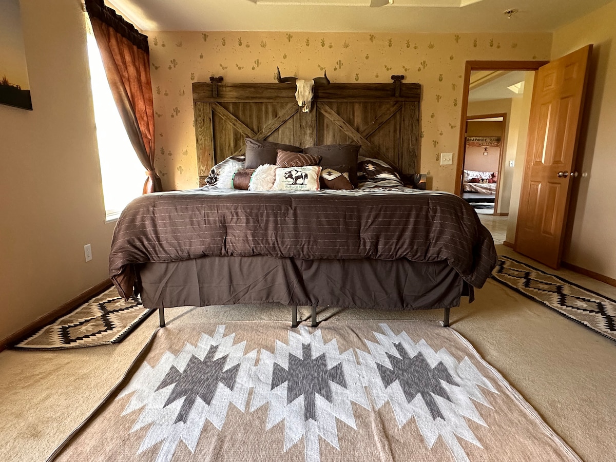 A warm sleeping area features a large, dark-colored bed with decorative pillows. The headboard, crafted from wood, includes rustic details. Peach walls and patterned rugs add to the room's character, while natural light enters through a window draped with rich curtains.
