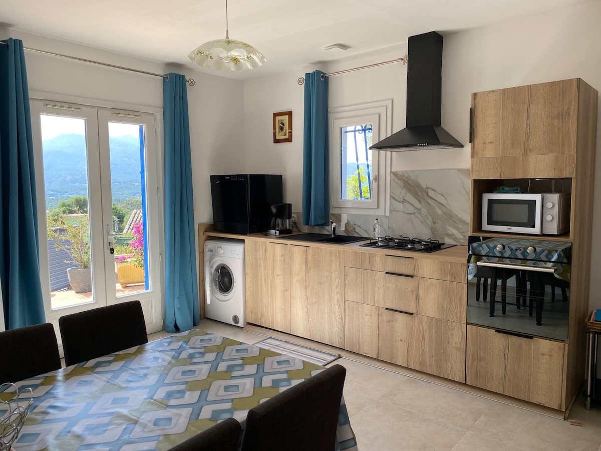 A modern kitchen area is presented with wooden cabinetry and stainless steel appliances. A dining table is set for several people. A window offers a view of the mountains, while a washer is positioned near the kitchen. Natural light fills the space through the open glass doors.