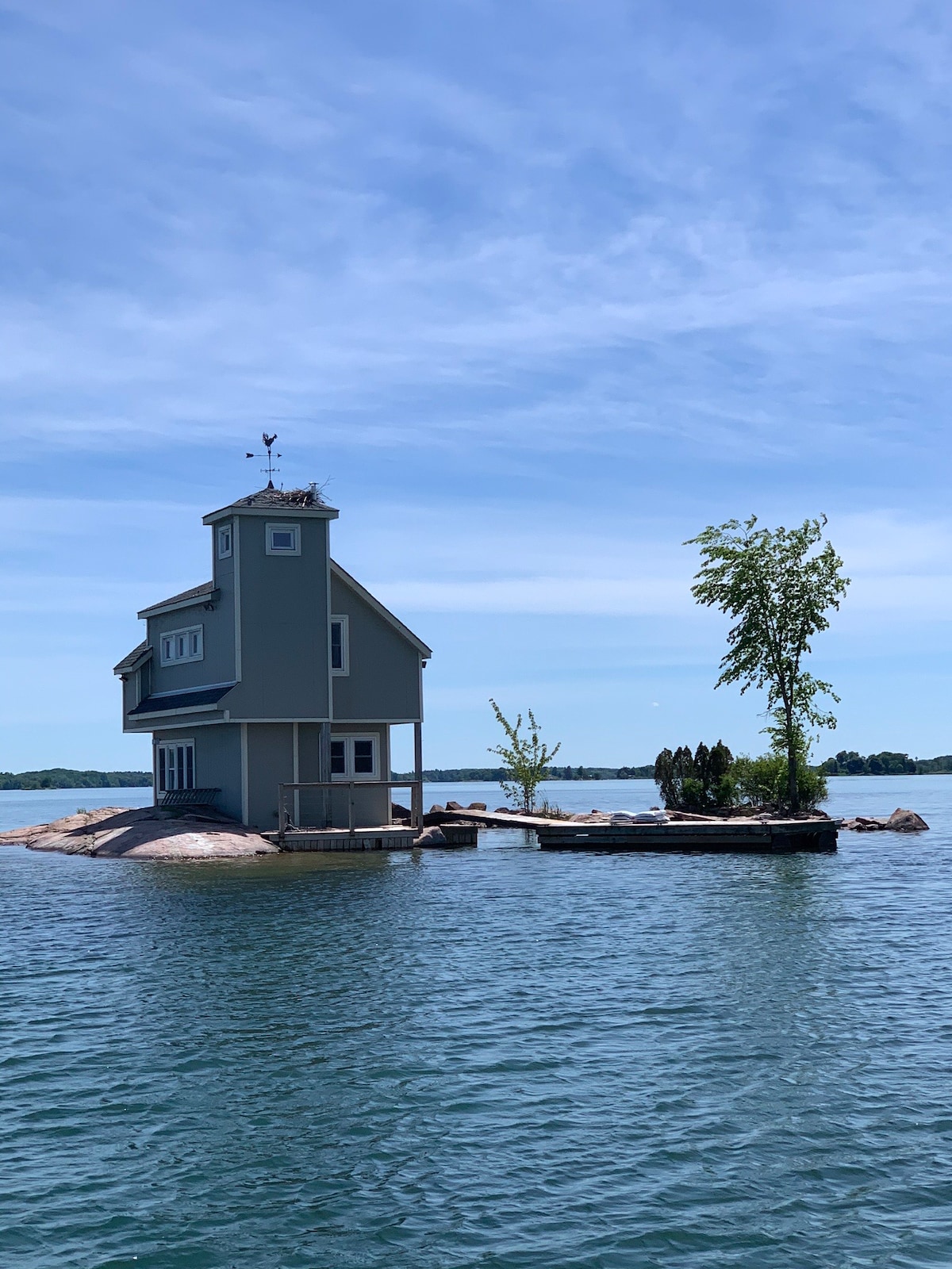 A quaint cottage is situated on a rocky island, surrounded by calm water. The structure features a light blue exterior and a simple porch, with trees surrounding the base. The peaceful landscape is enhanced by a clear blue sky.