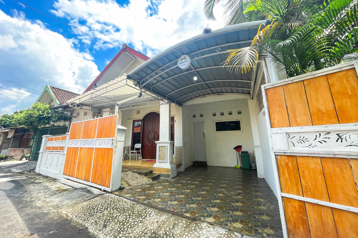 The exterior of the homestay is presented with a covered entrance and driveway, showcasing a blend of light-colored walls and wooden accents. A tiled pathway leads up to the entrance, while tropical plants add greenery to the welcoming atmosphere.