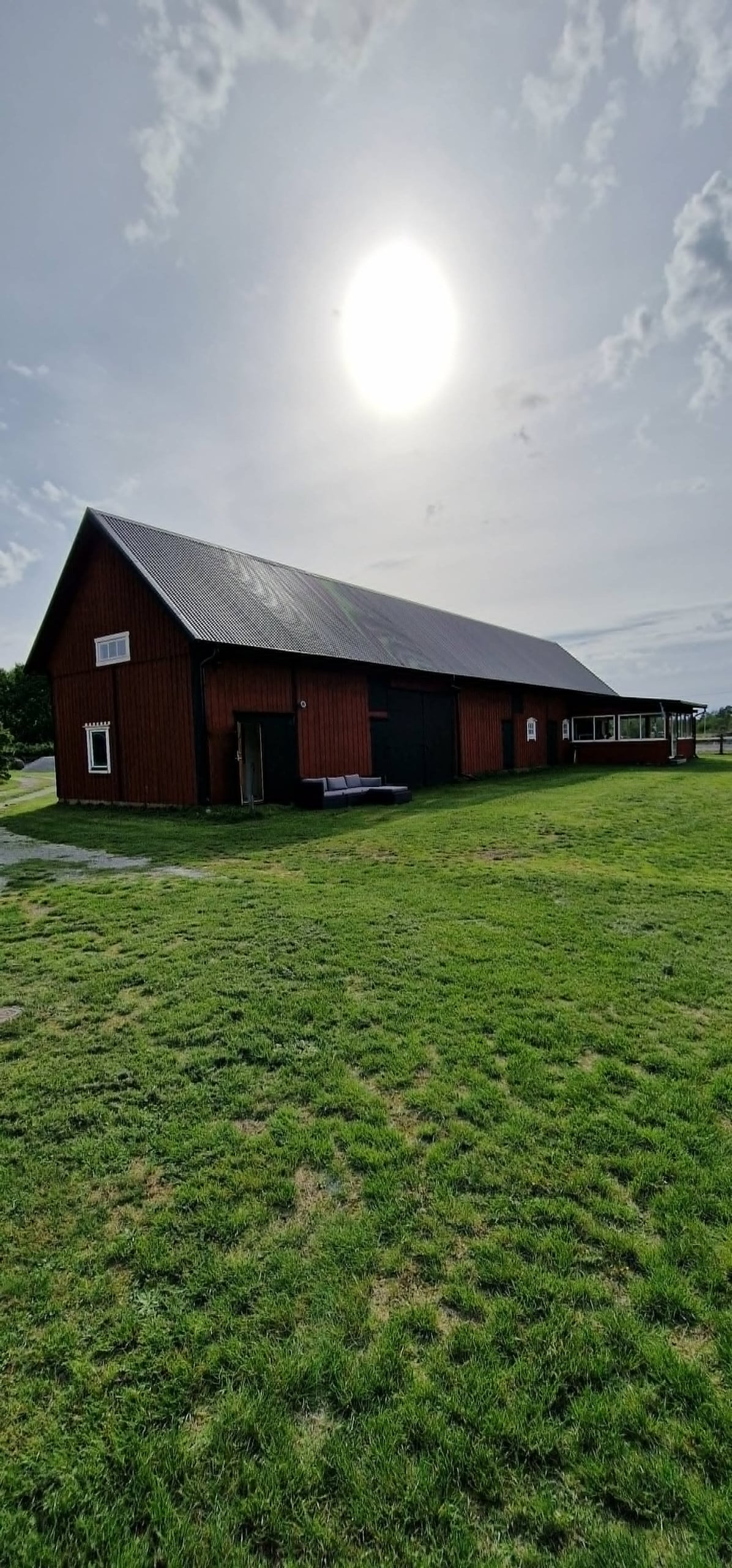 A large red barn structure is shown, featuring a sloped roof and open doors. Grass covers the surrounding area, with a clear sky overhead partially obscured by light clouds. Sunlight illuminates the scene, creating a natural brightness.