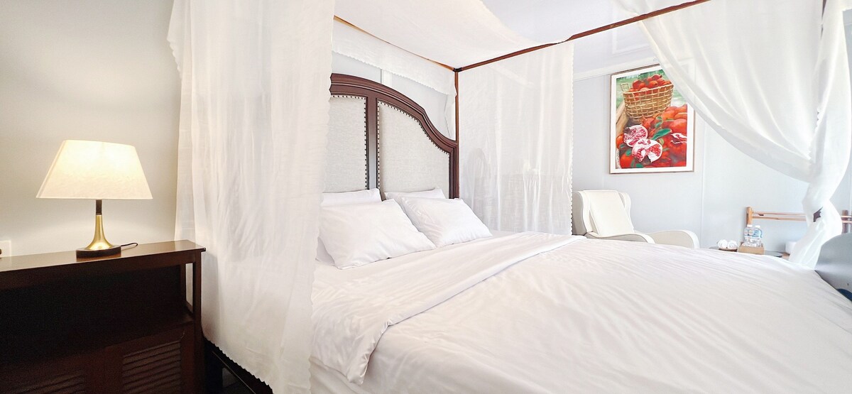 A spacious bed with white linens is set within a canopied frame, complemented by soft drapes. A bedside table with a lamp is present, along with an elegant chair and decorative wall art featuring fruits in vibrant colors.