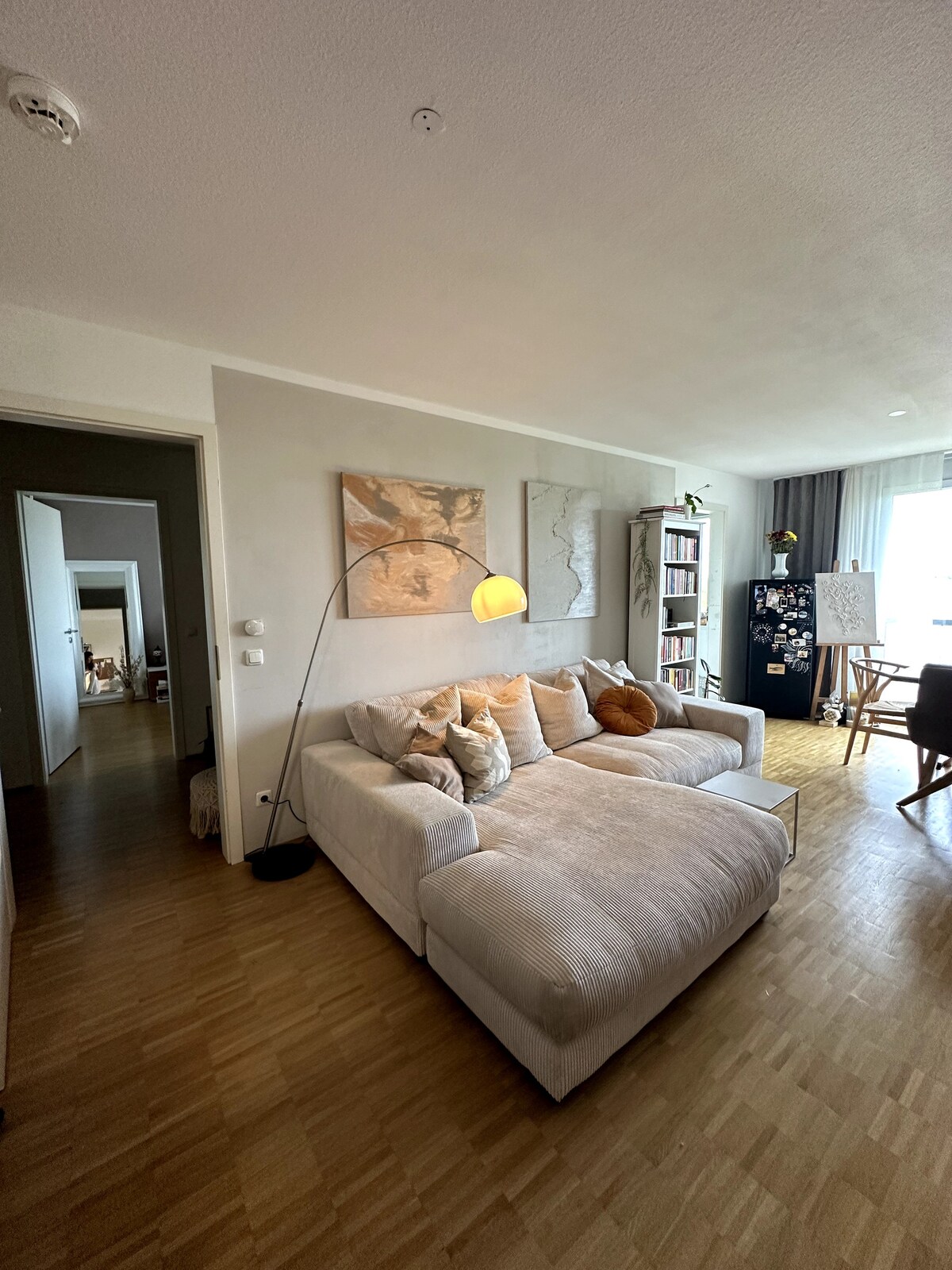 A light-filled living area is depicted, featuring a spacious sectional sofa adorned with an array of cushions. A contemporary floor lamp stands nearby, and a bookshelf filled with books is visible. Neutral tones dominate the decor, contributing to a calming environment.