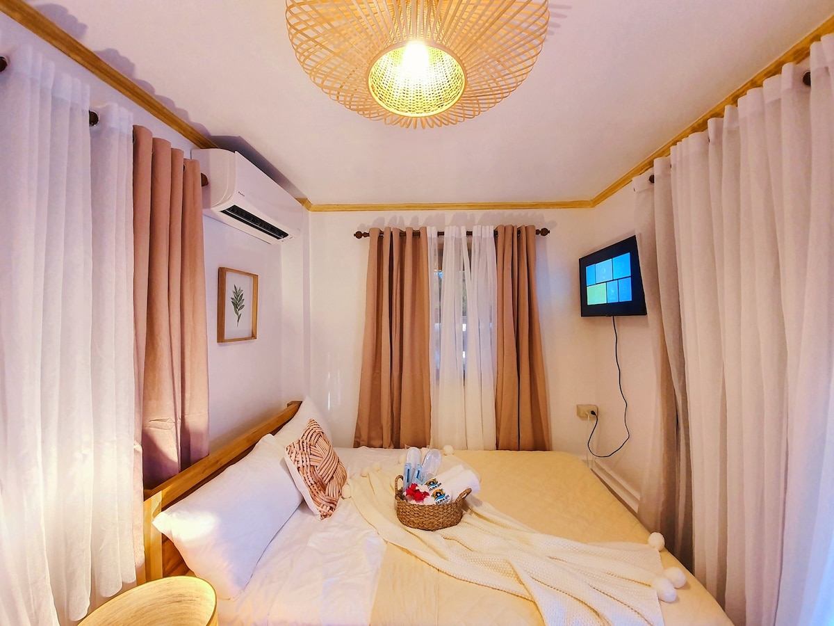 A cozy bedroom is presented with soft, neutral-toned curtains framing the windows. A spacious bed, adorned with a neatly arranged blanket, is complemented by a small decorative basket of towels. A ceiling light fixture provides gentle illumination, while a wall-mounted television is visible.