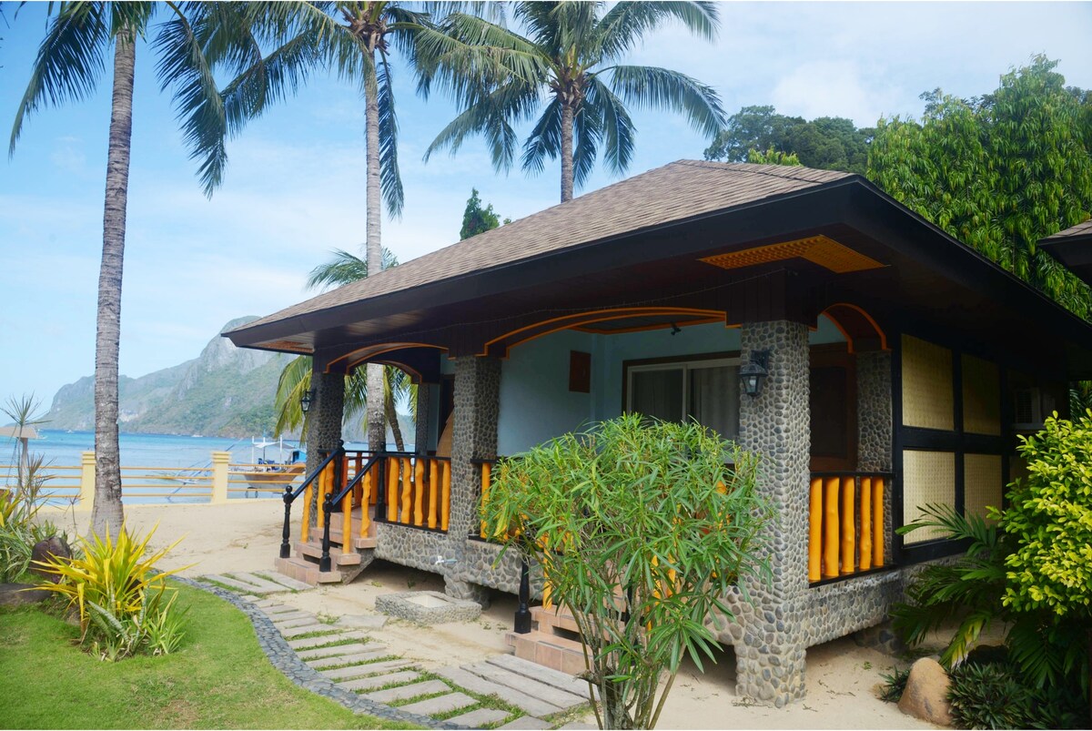 A beachfront villa is presented, featuring a wide terrace with wooden railings. Tropical palm trees and lush greenery surround the property, while a pathway leads to the entrance. The sea can be glimpsed beyond the villa, complementing its serene setting.