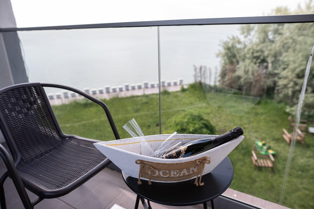 A balcony scene features a black chair beside a small table holding a decorative boat filled with champagne and two flutes. Views of lush greenery and the lake are visible in the background, creating a serene outdoor space.