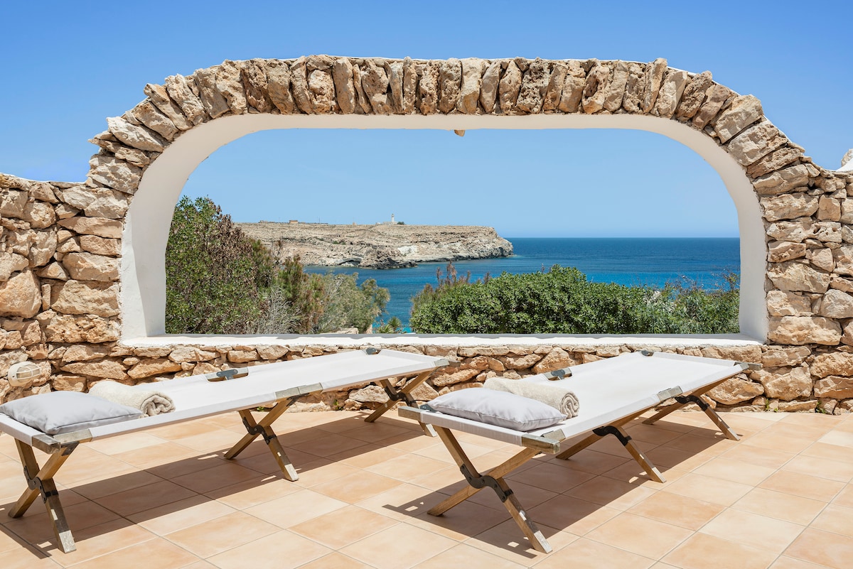 A serene terrace area features two sun loungers with soft cushions, positioned under a stone arch. The view highlights the expansive sea and the distant coastline, blending natural beauty and architectural detail.