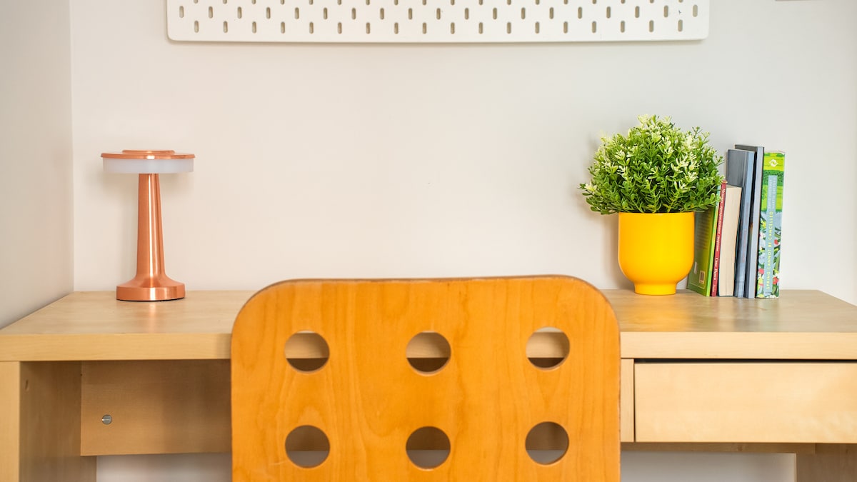 A modern workspace features a wooden desk with a light-colored finish, complemented by a chair with circular cut-outs. A copper accent lamp provides soft lighting, while a yellow planter with greenery sits beside a collection of books arranged neatly.